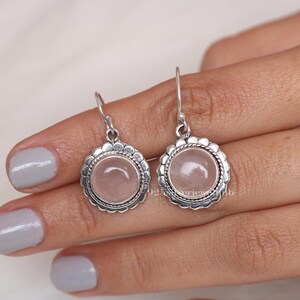 May include: A pair of round drop earrings featuring a smooth, pale pink rose quartz cabochon set in a sterling silver bezel. The circular setting is framed by a decorative scalloped edge and a twisted rope detail, suspended from silver hooks.