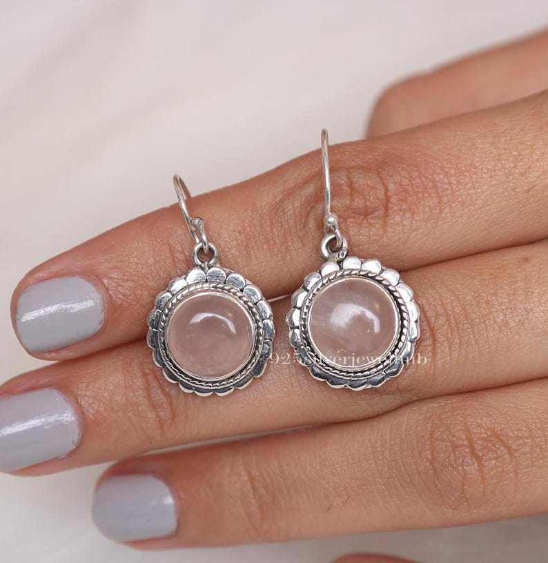 May include: A pair of round drop earrings featuring a smooth, pale pink rose quartz cabochon set in a sterling silver bezel. The circular setting is framed by a decorative scalloped edge and a twisted rope detail, suspended from silver hooks.