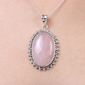 May include: A silver pendant necklace featuring a large, smooth, oval-shaped rose quartz gemstone. The stone is set in a decorative silver frame with a beaded edge, suspended from a delicate silver chain that rests against the skin.
