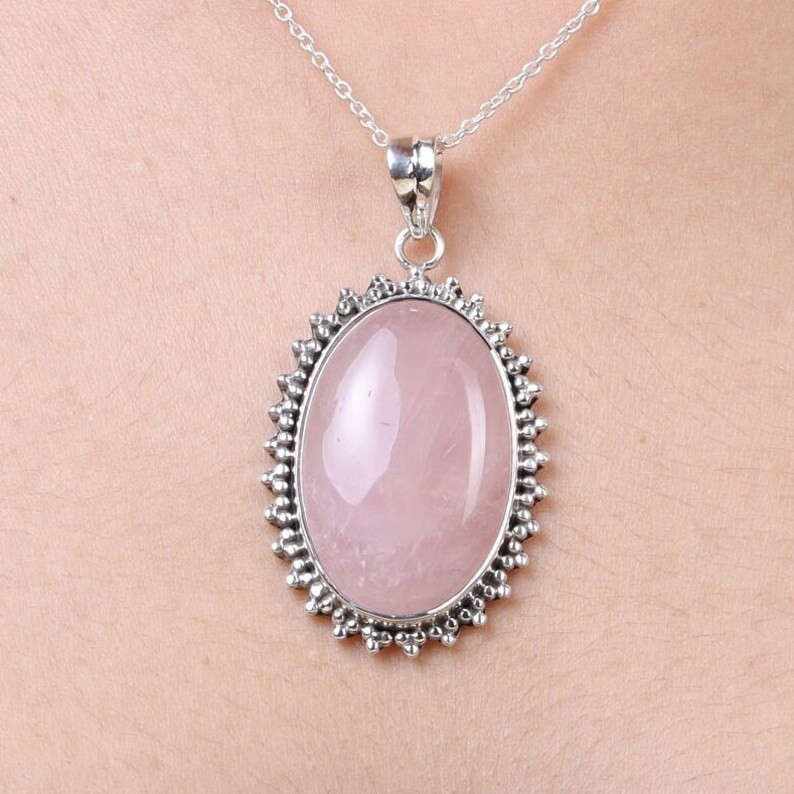 May include: A silver pendant necklace featuring a large, smooth, oval-shaped rose quartz gemstone. The stone is set in a decorative silver frame with a beaded edge, suspended from a delicate silver chain that rests against the skin.