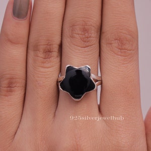 May include: A sterling silver ring featuring a black star-shaped stone. The ring has a split band design and a polished finish. The image shows the ring on a hand, highlighting the contrast between the silver and the dark stone.