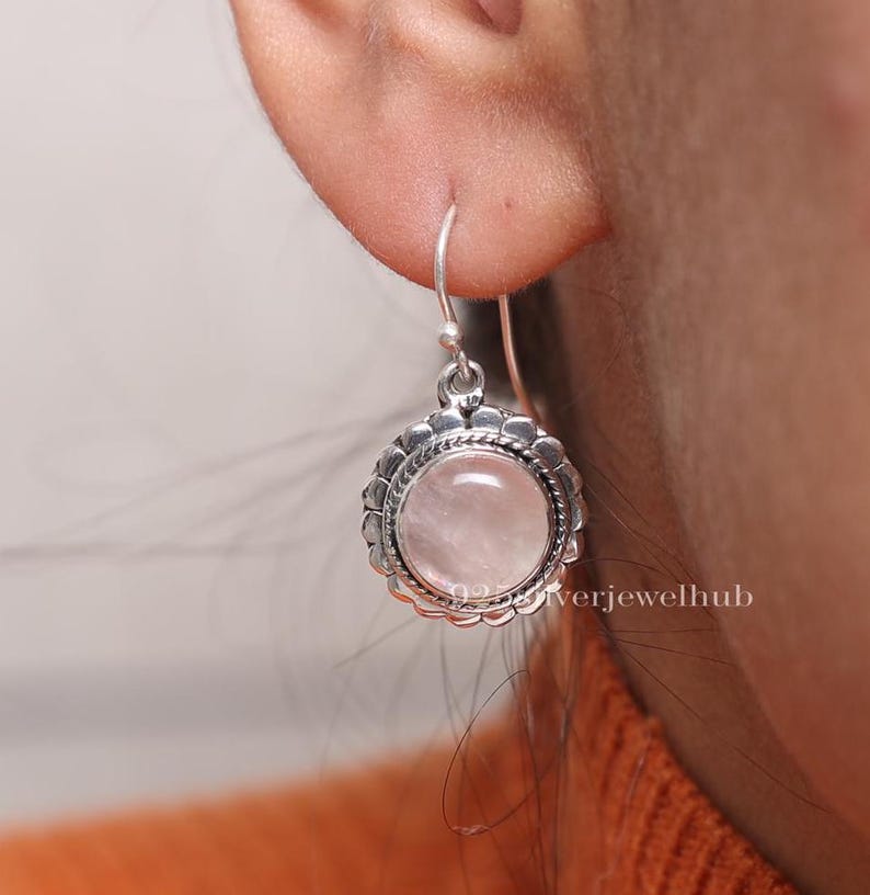 May include: A round earring featuring a smooth, pale pink rose quartz cabochon set in a sterling silver frame with a scalloped edge and a twisted rope detail. The earring hangs from a simple silver hook wire, displaying a polished finish.