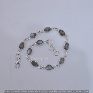 May include: A silver chain bracelet featuring eight oval-shaped labradorite gemstones with iridescent blue flashes. The bracelet includes a lobster claw clasp and a short extender chain with circular links, showcasing a polished metallic finish.
