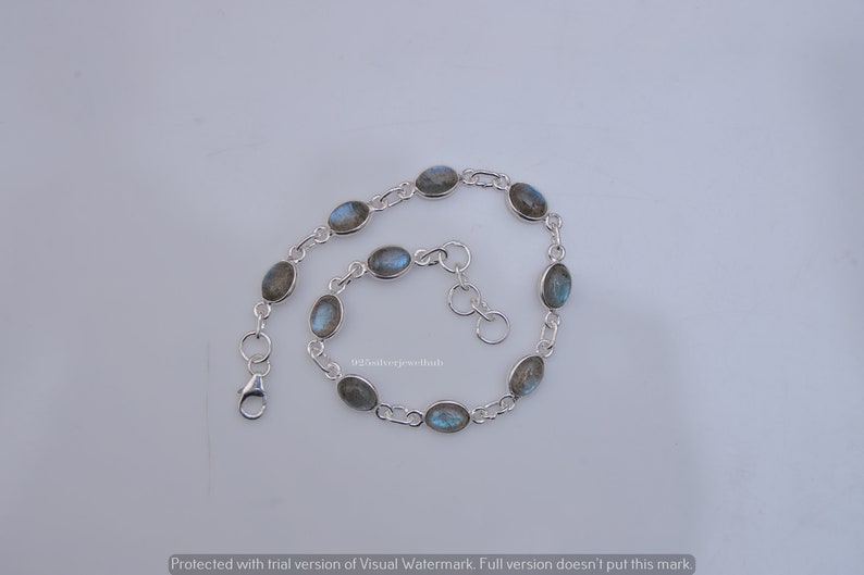 May include: A silver chain bracelet featuring eight oval-shaped labradorite gemstones with iridescent blue flashes. The bracelet includes a lobster claw clasp and a short extender chain with circular links, showcasing a polished metallic finish.