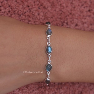 May include: A silver chain bracelet featuring five oval-shaped, iridescent grey-blue labradorite gemstones set in smooth metal bezels. The bracelet is linked with small silver rings and includes the text 925silverjewelhub printed on the side.