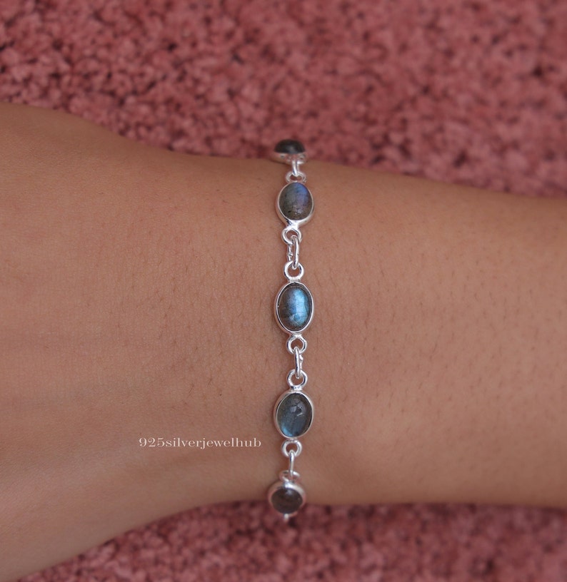 May include: A silver chain bracelet featuring five oval-shaped, iridescent grey-blue labradorite gemstones set in smooth metal bezels. The bracelet is linked with small silver rings and includes the text 925silverjewelhub printed on the side.