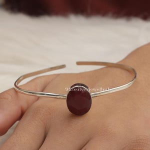 May include: A thin silver cuff bracelet featuring a single, central, oval-shaped, deep red faceted gemstone. The metal band is smooth and polished, with an open design. The text 925silverjewelhub is visible on the surface of the red stone.