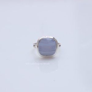 May include: A silver ring featuring a smooth, cushion-cut, light blue chalcedony gemstone with subtle horizontal banding. The metal band has a split design on the sides. The text 925silverjewelhub is printed in white below the ring.