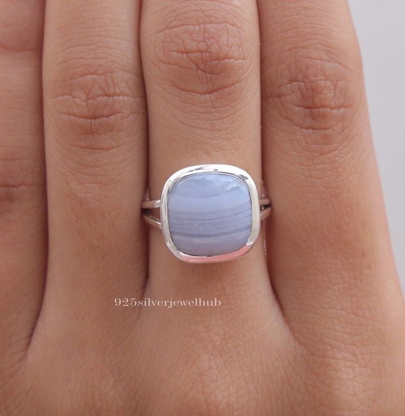 May include: A sterling silver ring featuring a smooth, cushion-cut blue lace agate gemstone with soft white banding. The metal band has a split design on the sides. The text 925silverjewelhub is visible on the surface near the base of the ring.