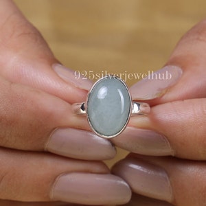 May include: A sterling silver ring featuring a smooth, oval-shaped, pale green cabochon gemstone set in a simple metal bezel. The text 925silverjewelhub is visible above the ring. The band is polished silver with a plain, rounded design.
