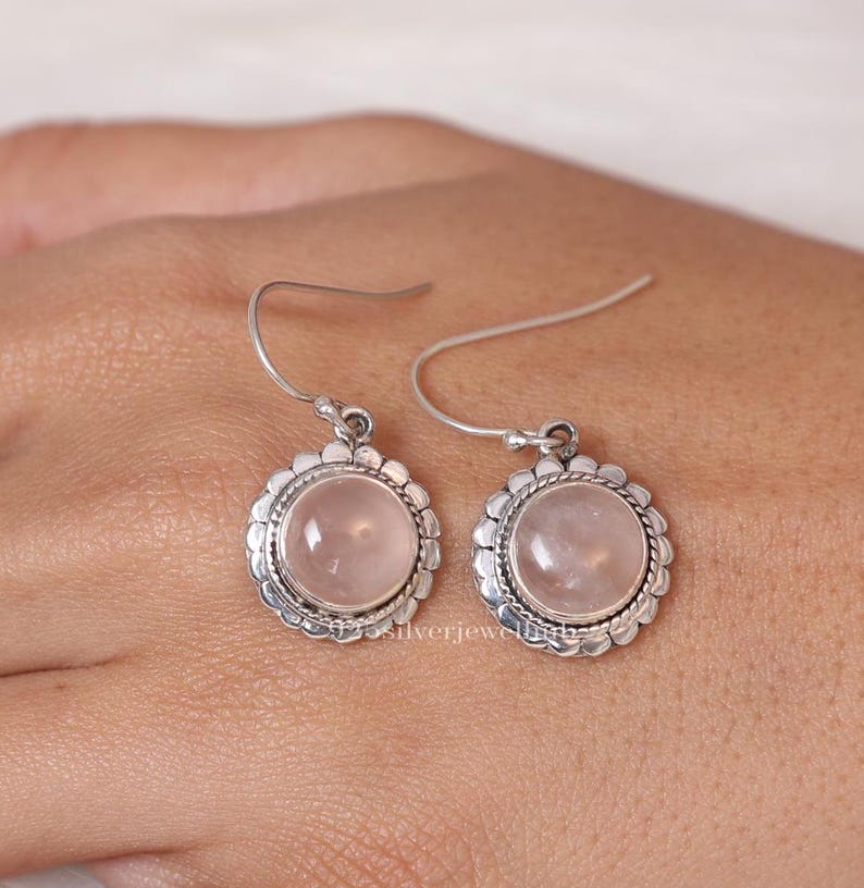 May include: A pair of dangle earrings featuring round, cabochon-cut pale pink rose quartz gemstones set in silver. Each stone is surrounded by a decorative scalloped silver border with a twisted rope detail. The earrings hang from simple silver hooks.