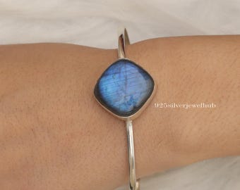 May include: A silver cuff bracelet featuring a central square-shaped labradorite gemstone with a blue iridescent sheen. The metal band is smooth and polished. The text 925silverjewelhub is visible on the right side of the gemstone setting.