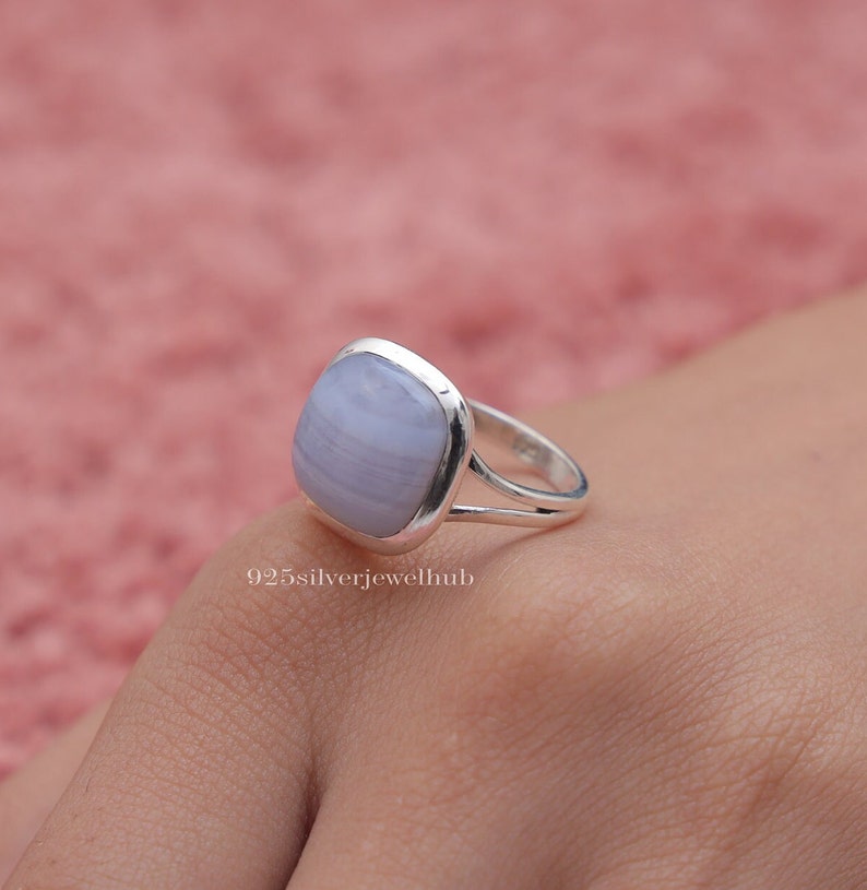 May include: A silver ring featuring a smooth, cushion-cut, light blue banded gemstone set in a polished metal bezel. The band has a split-shank design. The text 925silverjewelhub is visible on the surface below the ring. The ring is worn on a finger.