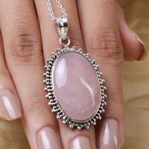 May include: An oval-shaped rose quartz pendant set in a decorative silver frame with a beaded edge. The pendant hangs from a delicate silver chain, showcasing a smooth, pale pink gemstone with natural internal inclusions and a polished finish.