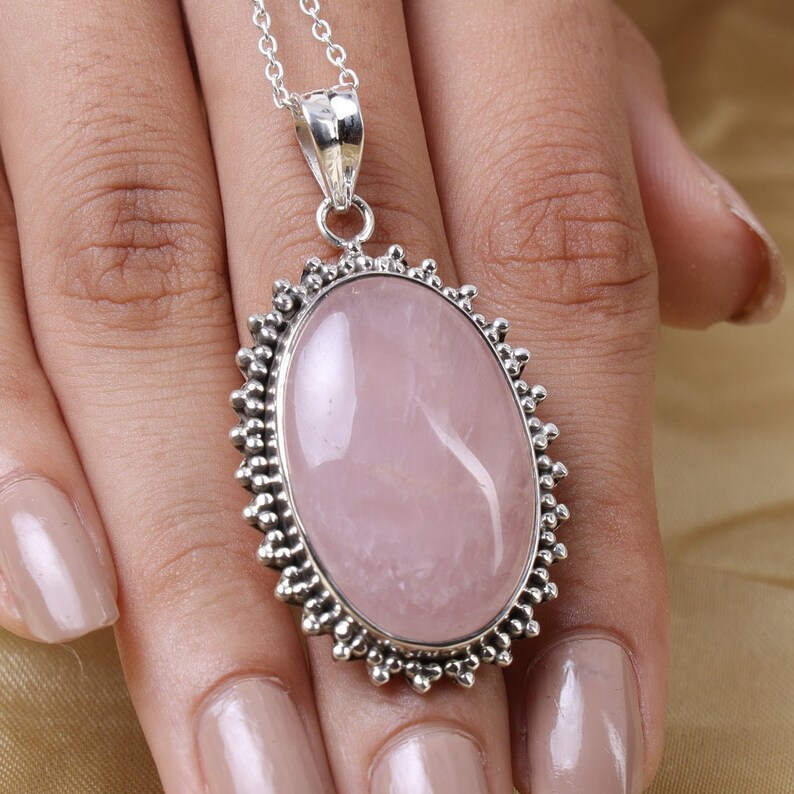 May include: An oval-shaped rose quartz pendant set in a decorative silver frame with a beaded edge. The pendant hangs from a delicate silver chain, showcasing a smooth, pale pink gemstone with natural internal inclusions and a polished finish.