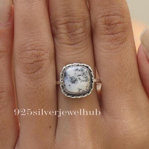 May include: A silver ring with a square-shaped white and black dendritic agate gemstone. The ring has a rope-like design around the edge of the setting.