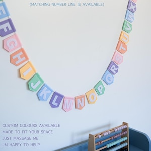 Puede incluir: Una guirnalda de letras del alfabeto de color arcoíris pastel con letras blancas cuelga de una pared blanca. Debajo, un ábaco de madera se encuentra en un estante azul con contenedores coloridos. El texto en la pared dice "PASTEL RAINBOW COLOUR".
