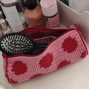 Puede incluir: Un neceser de ganchillo rosa y rojo con un estampado de lunares. La bolsa está abierta, revelando un cepillo para el pelo negro. La bolsa está sobre una superficie blanca, con varios productos de belleza en el fondo.