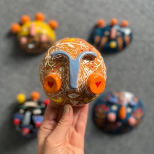 May include: Five ceramic wall masks with different designs. The masks are painted in various colors, including orange, yellow, blue, red, and white. The masks have different facial features, including eyes, noses, and mouths.
