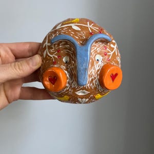 May include: A ceramic wall hanging in the shape of a face with a blue nose, orange cheeks, and white painted details. The face has red hearts on the cheeks and yellow birds on the top and bottom. The background is brown with white painted leaves and red berries.