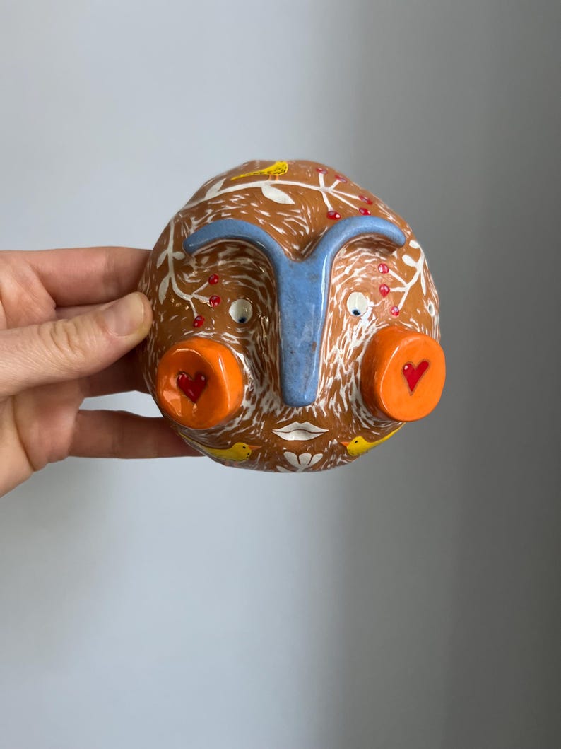 May include: A ceramic wall hanging in the shape of a face with a blue nose, orange cheeks, and white painted details. The face has red hearts on the cheeks and yellow birds on the top and bottom. The background is brown with white painted leaves and red berries.