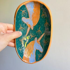 Può includere: Vassoio ovale in ceramica per bigiotteria con sfondo verde acqua e bordo dorato. Il vassoio presenta due unicorni blu con criniere e corna dorate, circondati da accenti floreali dorati. Il vassoio è tenuto in mano.