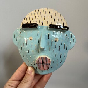 May include: A ceramic face sculpture with a light blue face, black eyebrows, and a pink and black mouth. The top of the head is cream-colored with black dashes. The sculpture is held by a hand.