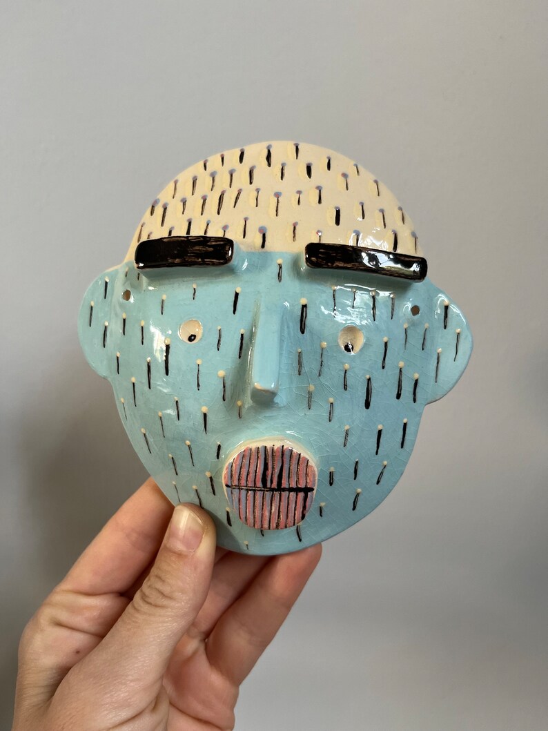 May include: A ceramic face sculpture with a light blue face, black eyebrows, and a pink and black mouth. The top of the head is cream-colored with black dashes. The sculpture is held by a hand.