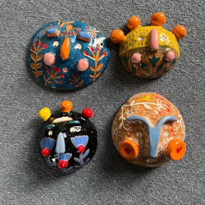 May include: Four ceramic wall hangings with whimsical faces. Each face is painted with different colors and patterns, including blue, orange, yellow, green, red, and black. The faces have unique features, such as a crown, a nose, and eyes.