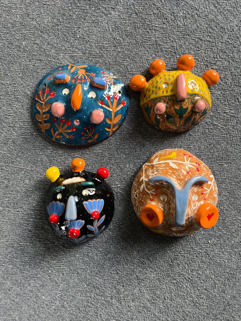 May include: Four ceramic wall hangings with whimsical faces. Each face is painted with different colors and patterns, including blue, orange, yellow, green, red, and black. The faces have unique features, such as a crown, a nose, and eyes.