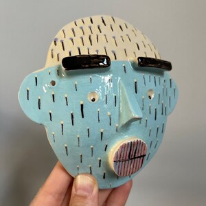 May include: A ceramic face sculpture with a light blue face and a cream-colored top. The face has black eyebrows, small eyes, a nose, and a mouth with pink and black stripes. The face is decorated with black and yellow dashes.