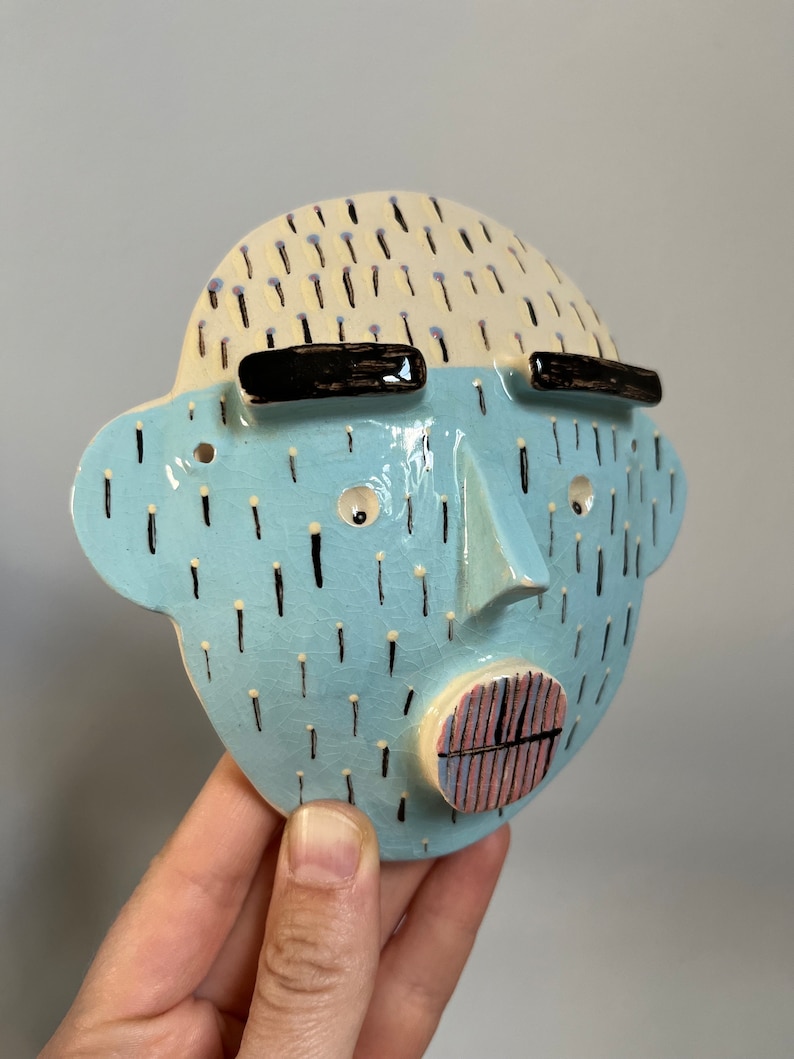 May include: A ceramic face sculpture with a light blue face and a cream-colored top. The face has black eyebrows, small eyes, a nose, and a mouth with pink and black stripes. The face is decorated with black and yellow dashes.