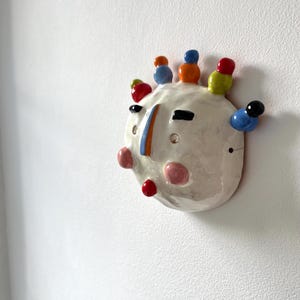 Op de afbeelding: Een keramische wandsculptuur in de vorm van een gezicht. Het gebroken witte gezicht heeft kleurrijke accenten, waaronder rode, blauwe, oranje en groene details. De sculptuur heeft een glanzende afwerking en is bevestigd aan een witte muur.