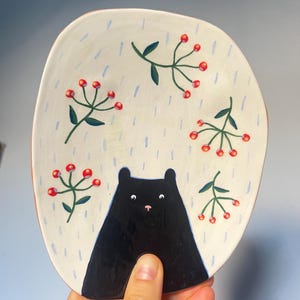 Könnte beinhalten: Handbemalte Keramikplatte mit einem Design eines schwarzen Bären. Die cremefarbene Platte zeigt eine schwarze Bärensilhouette, rote Beeren und grüne Blätter. Blaue, regenartige Linien sind auf die Platte gemalt. Die Platte wird von einer Hand gehalten.