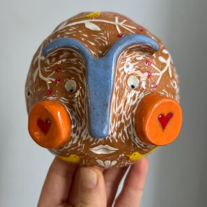 May include: A ceramic face sculpture with a brown base, white and red accents, and blue nose. The face has large orange eyes with red hearts, a white mouth, and yellow birds on the sides.