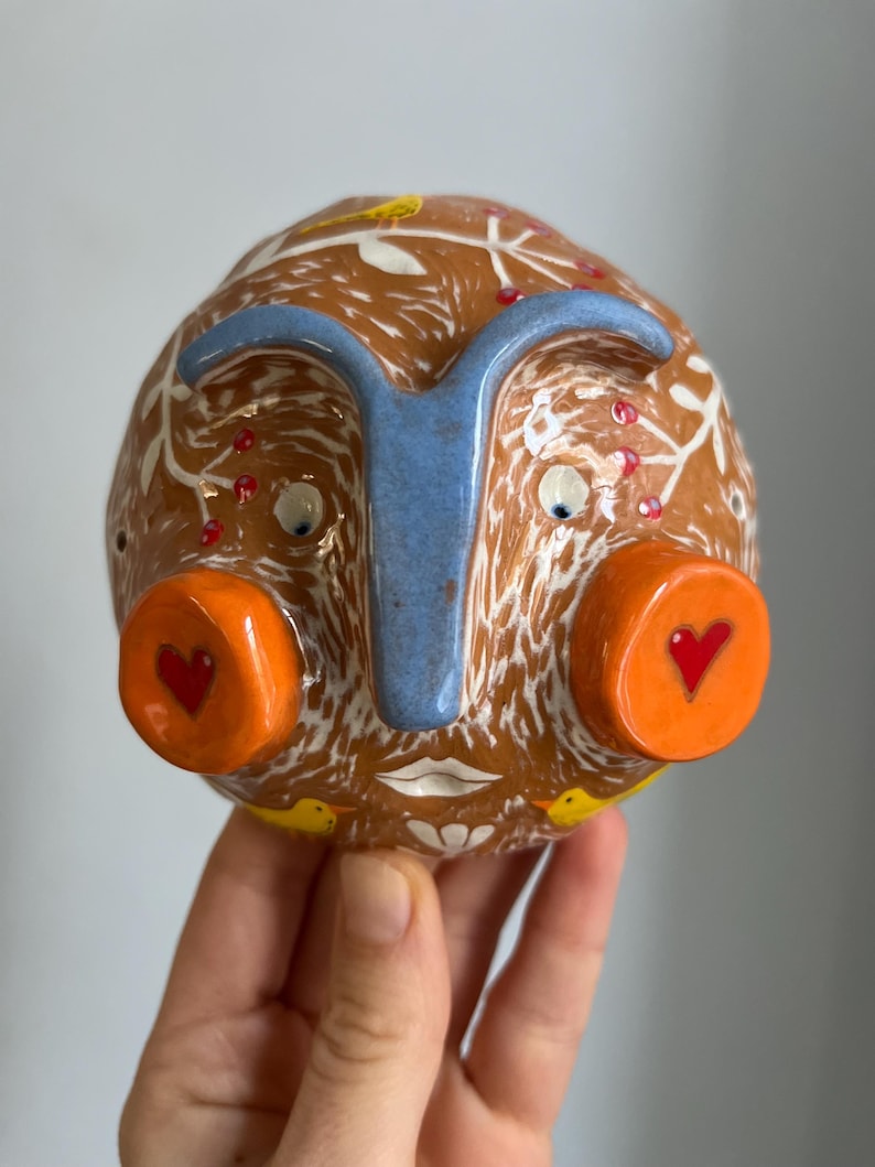 May include: A ceramic face sculpture with a brown base, white and red accents, and blue nose. The face has large orange eyes with red hearts, a white mouth, and yellow birds on the sides.
