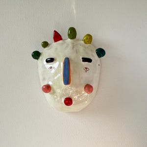 Könnte beinhalten: Eine skurrile Keramikmaske mit cremefarbenem Grund. Sie hat eine blaue und orangefarbene Nase, schwarze Augenbrauen und rote und rosa Akzente. Die Maske ist mit bunten, abgerundeten Vorsprüngen verziert.