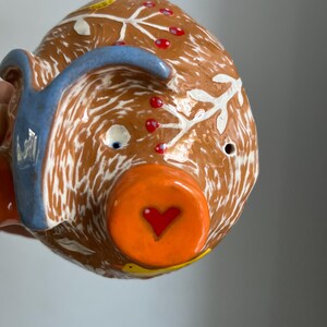 May include: A brown ceramic piggy bank with white and blue painted details. The piggy bank has a heart-shaped opening on the front and a small bird painted on the side.