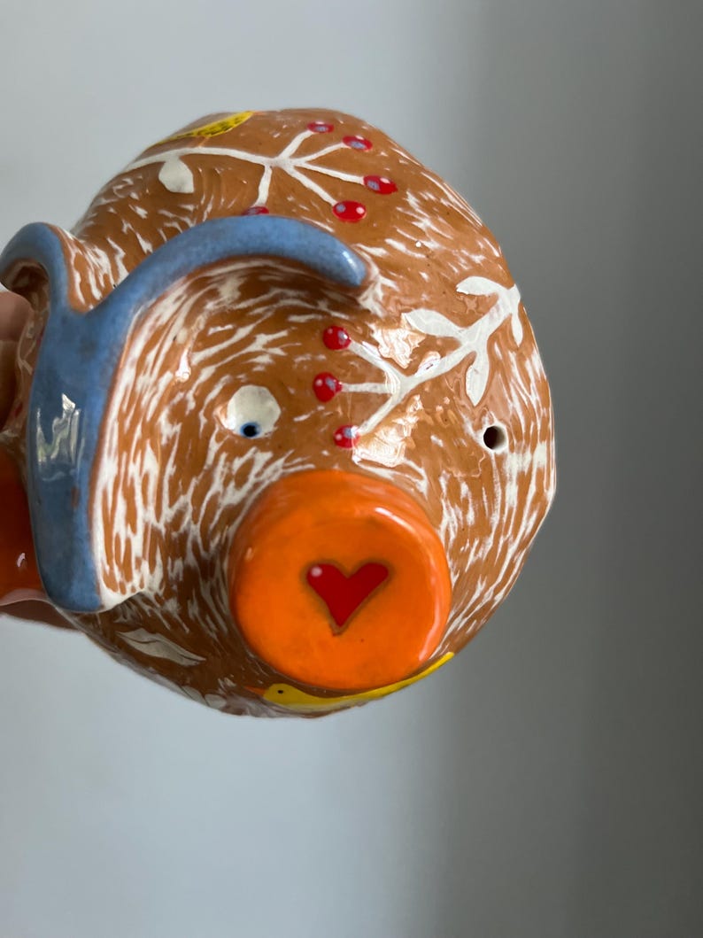 May include: A brown ceramic piggy bank with white and blue painted details. The piggy bank has a heart-shaped opening on the front and a small bird painted on the side.