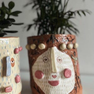 Puede incluir: Dos macetas de cerámica con caras pintadas a mano. La maceta de la izquierda tiene una cara azul claro, amarilla y rosa con una nariz azul. La maceta de la derecha tiene una cara blanca con una nariz y una boca rosas, y un fondo marrón, blanco y amarillo.