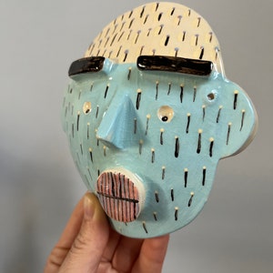 May include: A ceramic face sculpture with a light blue face, cream-colored hair, and black sunglasses. The face has a triangular nose, a pink and black mouth, and black and white details. The sculpture is held in a hand.