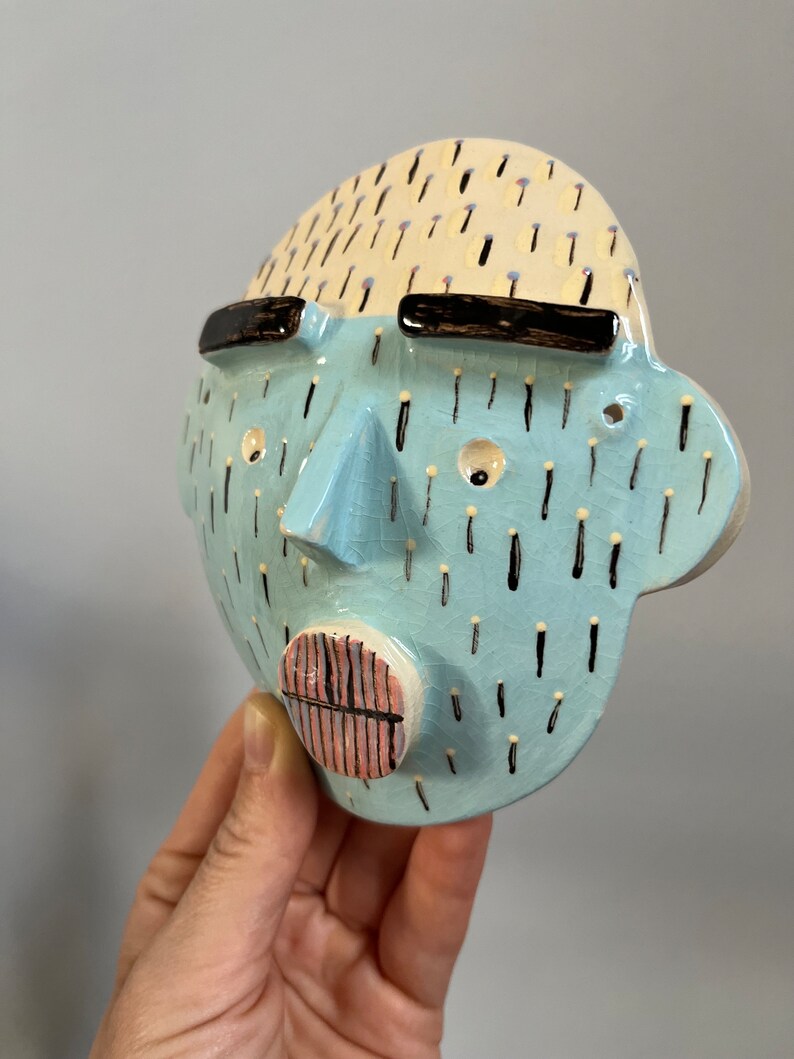 May include: A ceramic face sculpture with a light blue face, cream-colored hair, and black sunglasses. The face has a triangular nose, a pink and black mouth, and black and white details. The sculpture is held in a hand.
