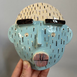 May include: A ceramic face mask with a light blue face, a cream-colored top, and black accents. The mask has black eyebrows, small eyes, and a mouth with pink and black stripes. The face is decorated with black and white vertical lines.