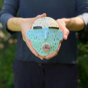 May include: A ceramic face sculpture with a light blue face, cream-colored hair, and black accents. The sculpture has a nose, mouth, and eyes. The face is held in front of a blurred background.