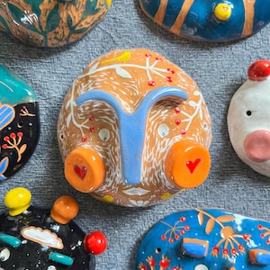 May include: A collection of colorful ceramic wall hangings with whimsical faces and floral designs. Each piece features unique details, including hearts, dots, and branches.