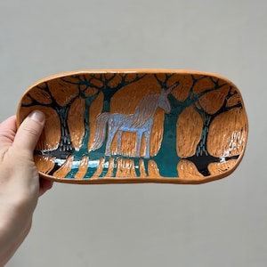 Può includere: Un vassoio rettangolare in ceramica con una scena boschiva stravagante. Il vassoio presenta un unicorno azzurro in una foresta turchese con alberi marroni. Il vassoio ha un bordo marrone.