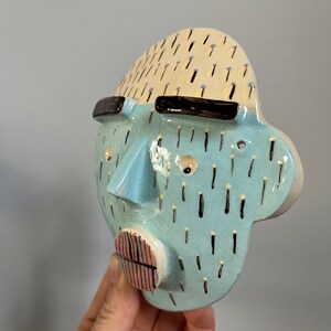 May include: A ceramic face sculpture with a light blue face, a beige top, and black accents. The sculpture has a nose, mouth, and eyes. The face is decorated with black dashes and is held by a hand.