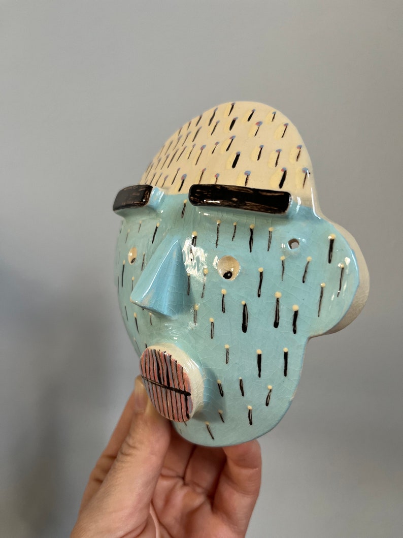 May include: A ceramic face sculpture with a light blue face, a beige top, and black accents. The sculpture has a nose, mouth, and eyes. The face is decorated with black dashes and is held by a hand.