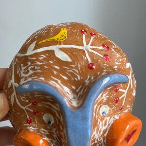 May include: A ceramic piggy bank with a brown and white glaze, a blue snout, and two orange ears. The piggy bank is decorated with a white branch with red berries and a yellow bird.