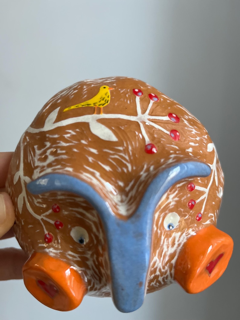 May include: A ceramic piggy bank with a brown and white glaze, a blue snout, and two orange ears. The piggy bank is decorated with a white branch with red berries and a yellow bird.
